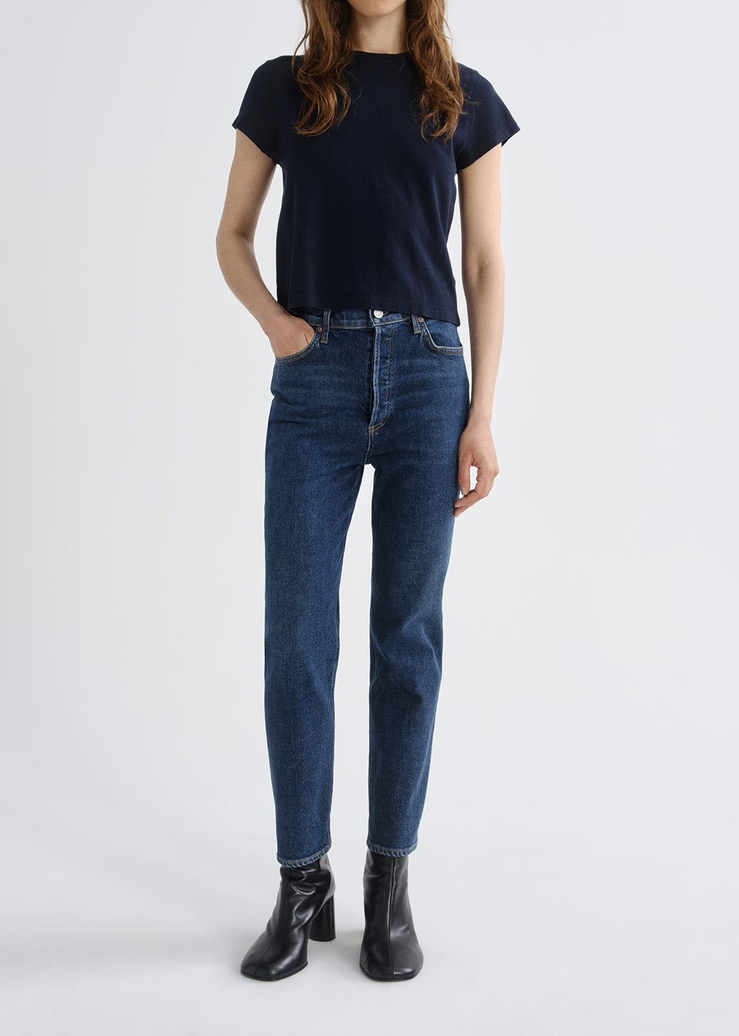 A model wearing a dark navy blue short-sleeve t-shirt, dark wash blue jeans, and black leather ankle boots, standing against a grey background.