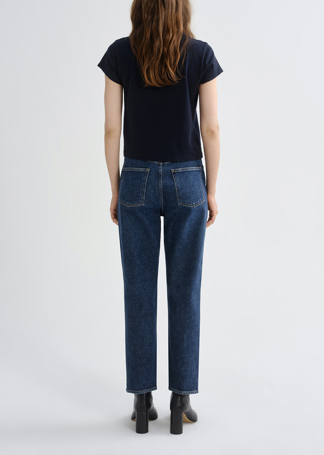 A model viewed from the back wearing dark blue straight-leg jeans, a dark navy t-shirt, and black ankle boots against a grey background.