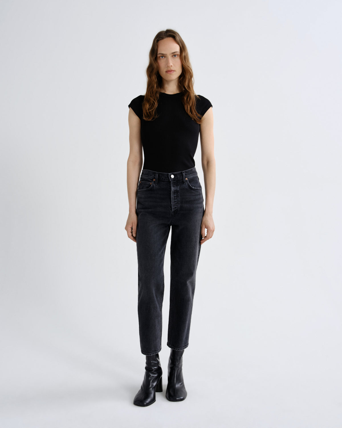 A woman stands against a light gray studio background wearing a fitted black short-sleeve top, dark charcoal high-waisted jeans, and black ankle boots.