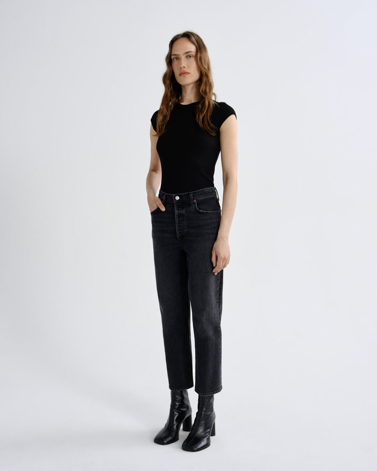 A woman stands against a light gray studio background wearing a fitted black short-sleeve top, dark charcoal jeans, and black ankle boots.