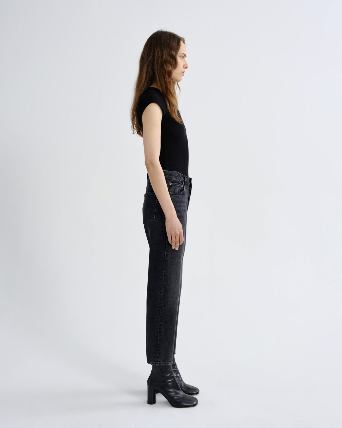 A woman stands in profile wearing a black sleeveless top, dark gray high-waisted straight-leg jeans, and black leather ankle boots against a plain light gray studio background.