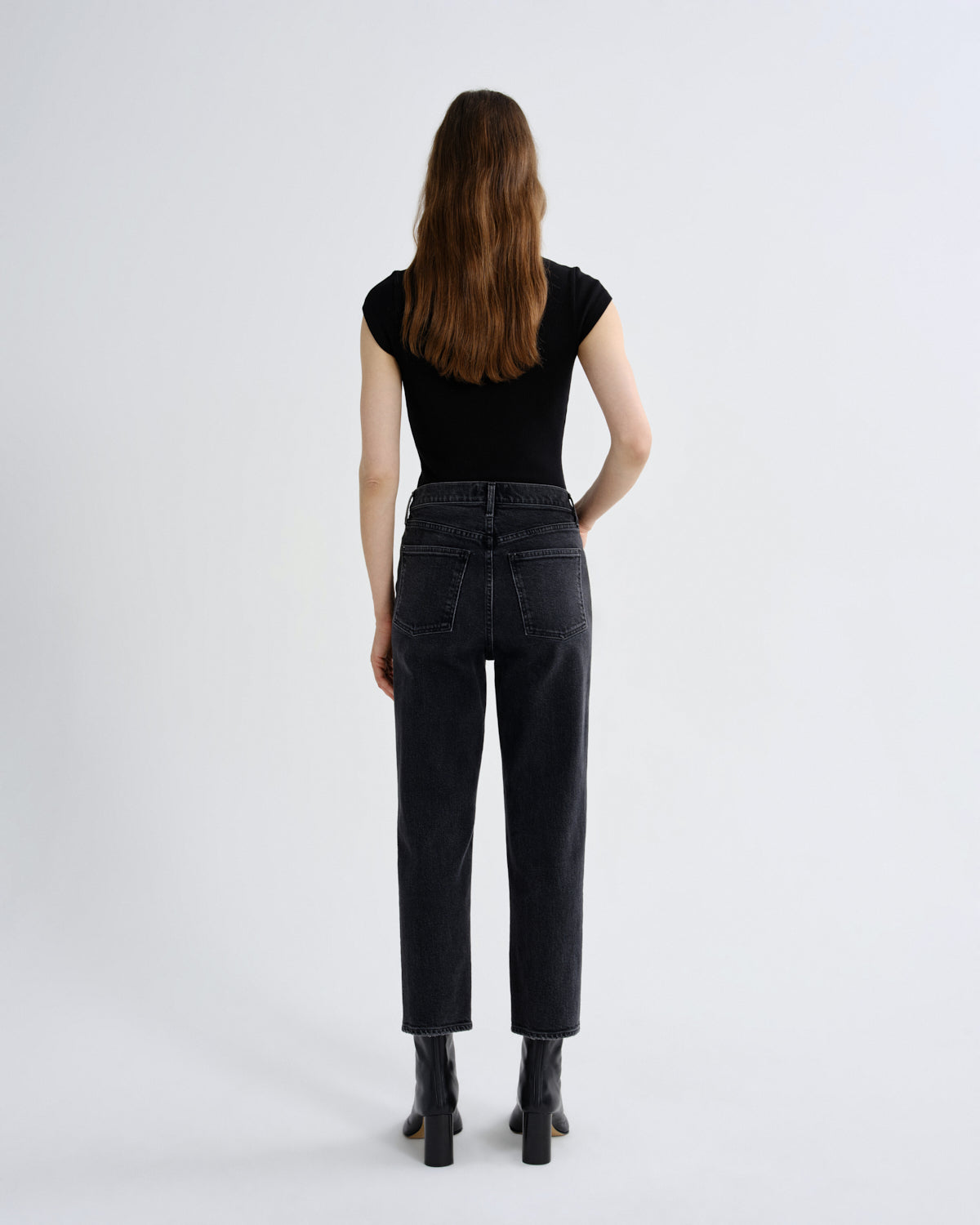 A woman viewed from behind wearing a black short-sleeve top, dark gray high-waisted straight-leg jeans, and black ankle boots, standing against a light gray studio background.