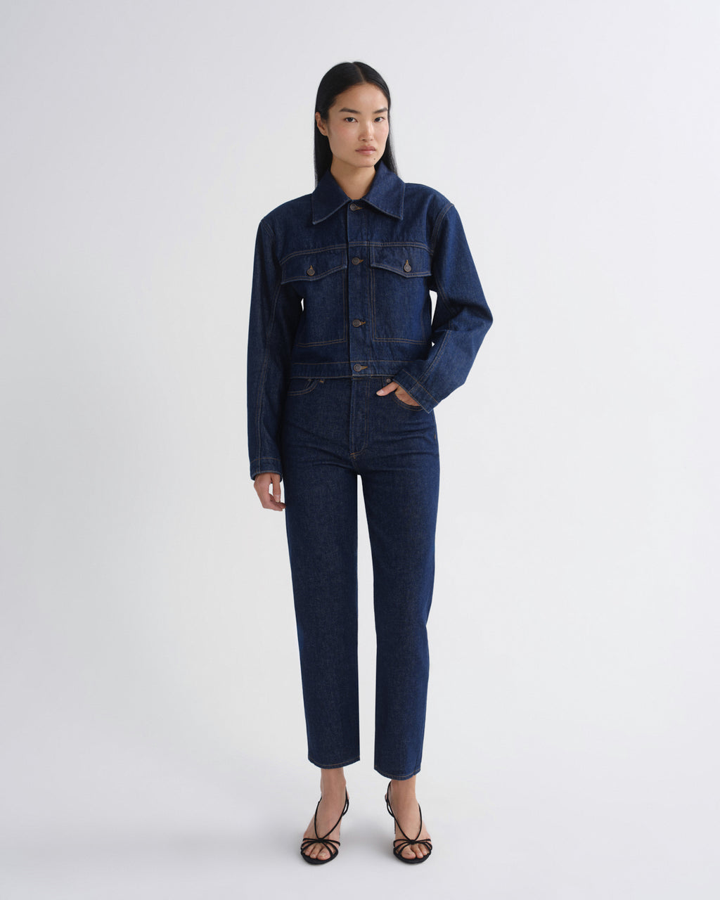 A model stands against a light gray studio background wearing a dark indigo denim jumpsuit with a button-up collar and front flap pockets, paired with black strappy heels.