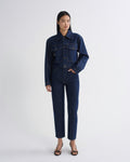 A model stands against a light gray studio background wearing a dark indigo denim jumpsuit with a button-up collar and front flap pockets, paired with black strappy heels.