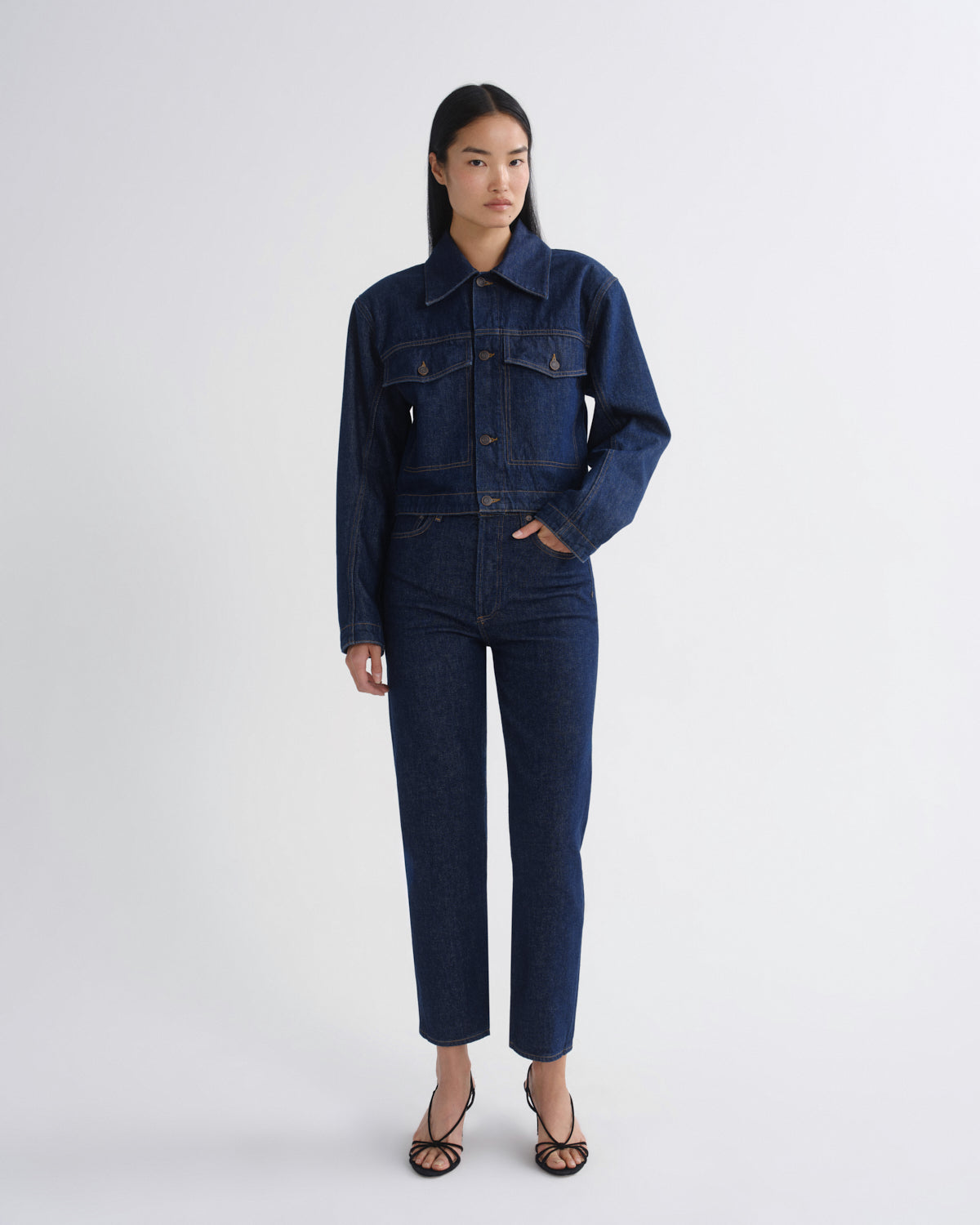 A model stands against a light gray studio background wearing a dark indigo denim jumpsuit with a button-up collar and front flap pockets, paired with black strappy heels.