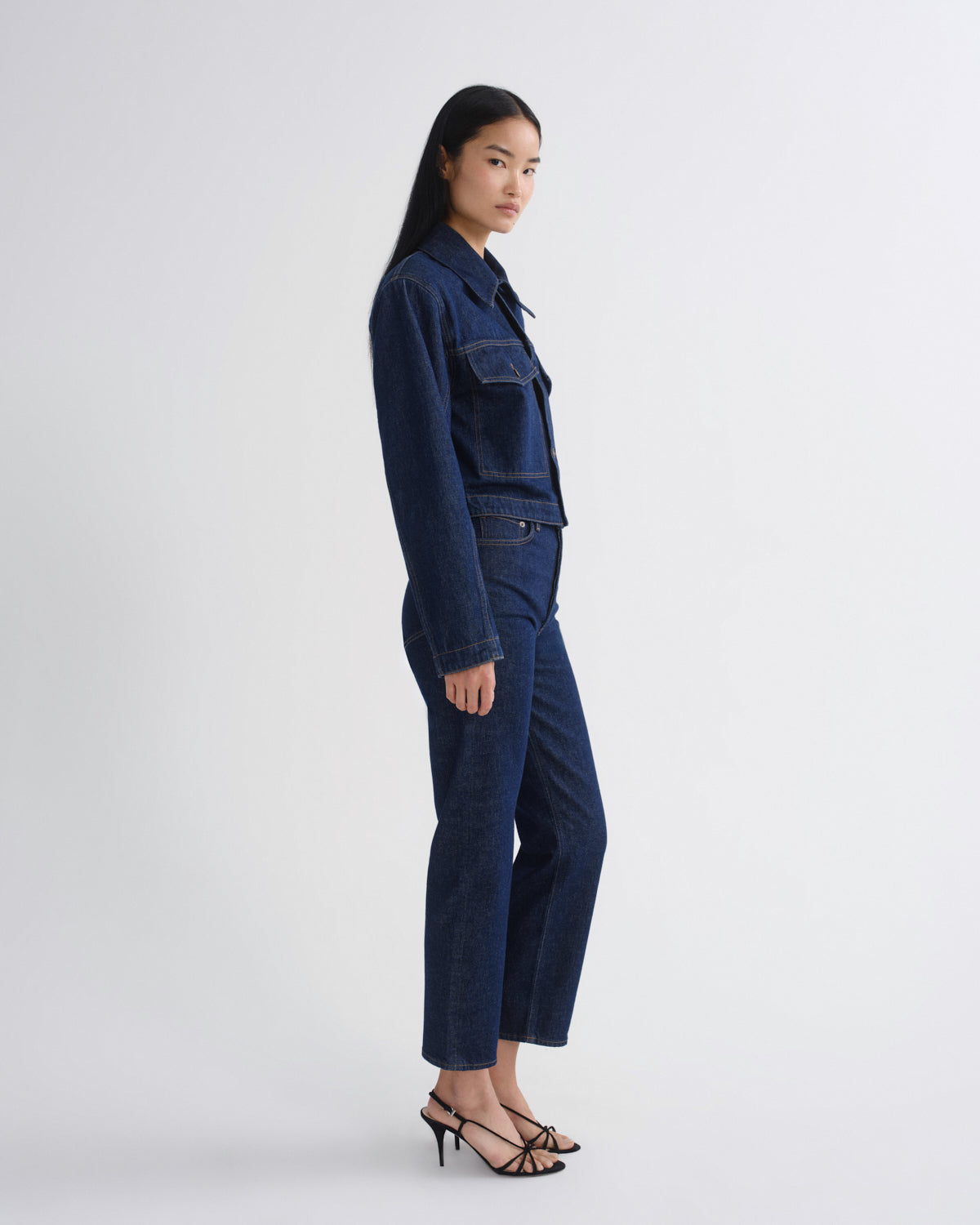 A model is shown in profile wearing a dark indigo denim set consisting of a cropped button-up jacket and matching cropped trousers, paired with black strappy high-heeled sandals, against a plain light gray studio background.