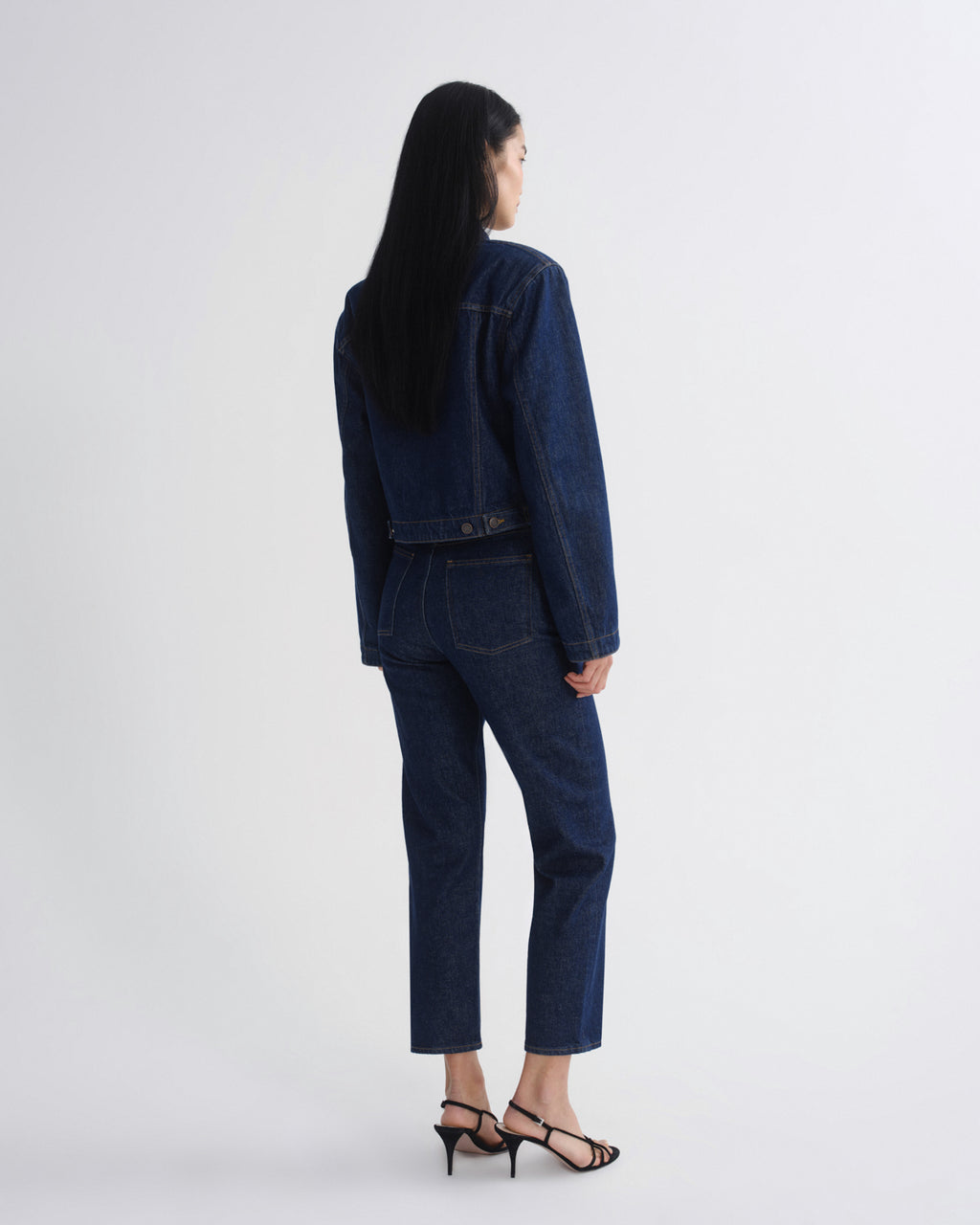 A model shown from the back wearing a dark blue denim jacket and matching cropped jeans, paired with black strappy heels, against a plain light gray studio background.
