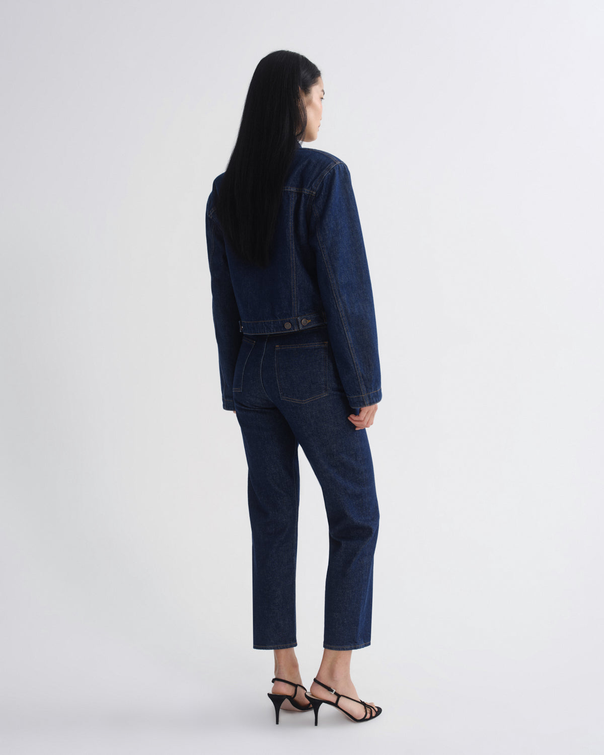 A model shown from the back wearing a dark blue denim jacket and matching cropped jeans, paired with black strappy heels, against a plain light gray studio background.