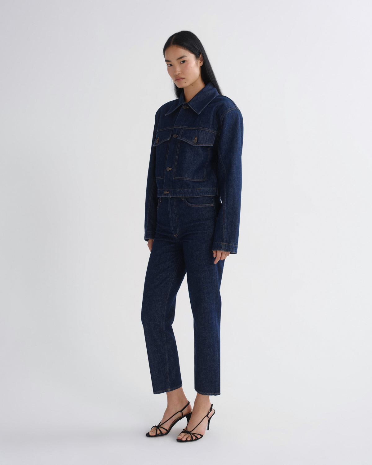 A model wearing a dark blue denim jumpsuit with a matching denim jacket and cropped pants, paired with black strappy high-heeled sandals, against a plain light gray studio background.