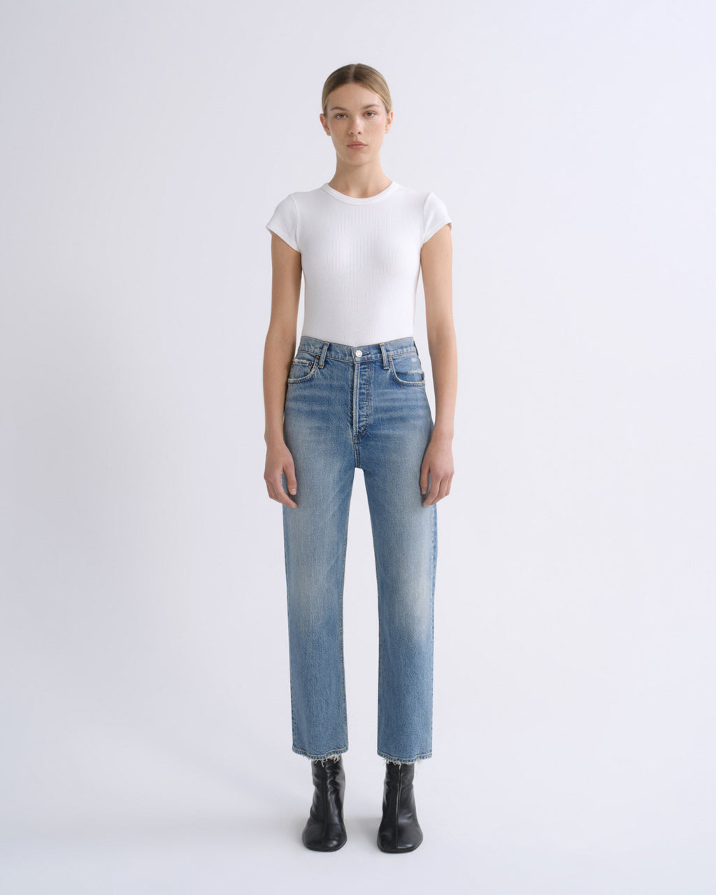A model stands against a light gray studio background wearing a fitted white short-sleeve top, light blue straight-leg jeans, and black ankle boots.