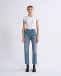 A model stands against a light gray studio background wearing a fitted white short-sleeve top, light blue straight-leg jeans, and black ankle boots.