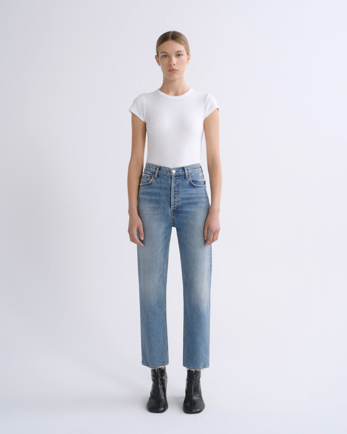 A model stands against a light gray studio background wearing a fitted white short-sleeve top, light blue straight-leg jeans, and black ankle boots.