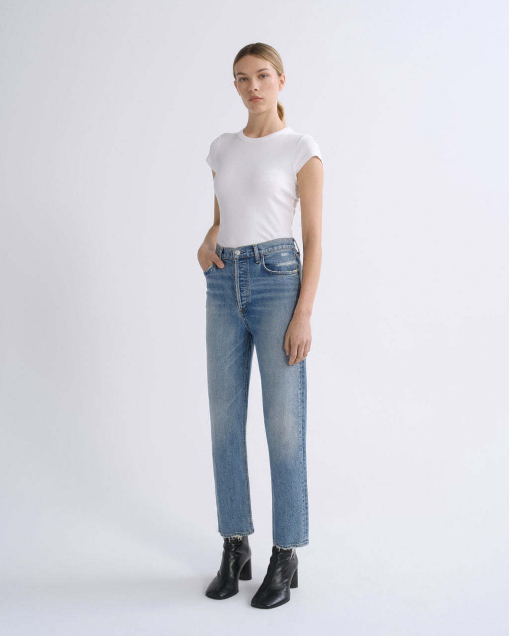 A woman modeling a pair of light-wash blue straight-leg jeans with a white short-sleeve crewneck t-shirt and black leather ankle boots, standing against a light gray studio background.