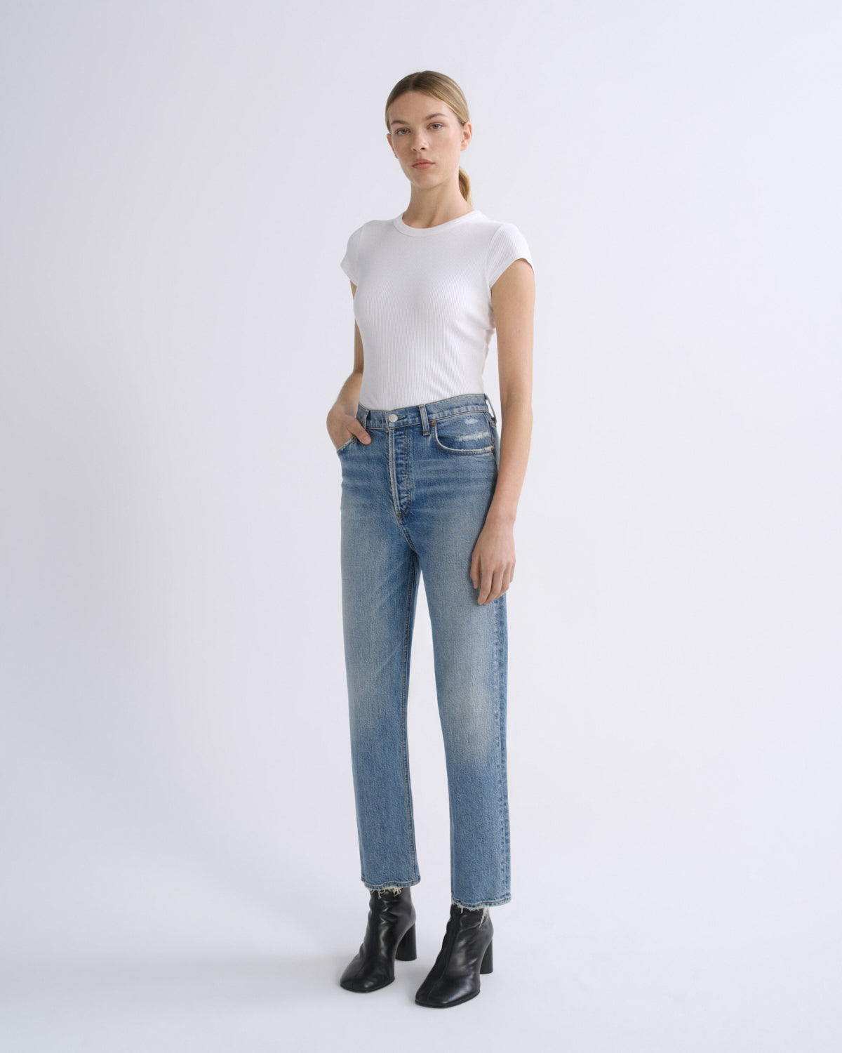 A woman modeling a pair of light-wash blue straight-leg jeans with a white short-sleeve crewneck t-shirt and black leather ankle boots, standing against a light gray studio background.