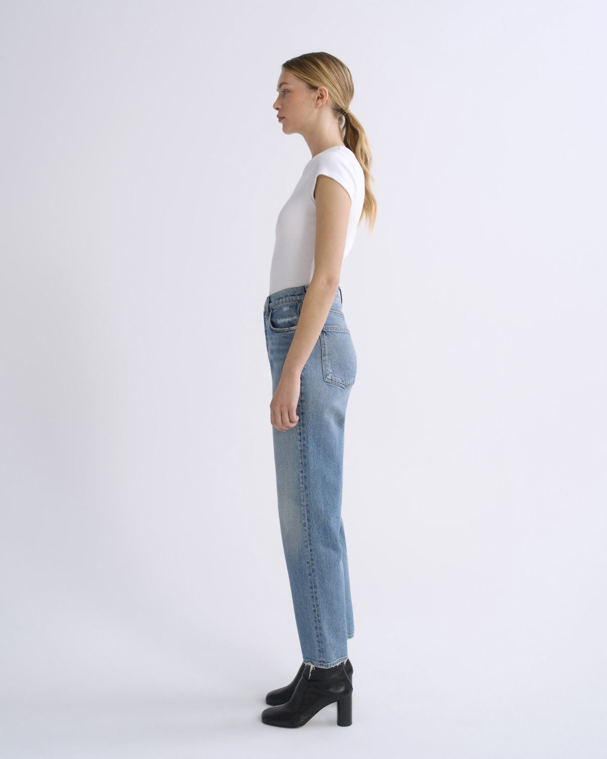 A woman in a side profile view wearing a white short-sleeve top, light-wash blue jeans, and black ankle boots, standing against a plain white studio background.
