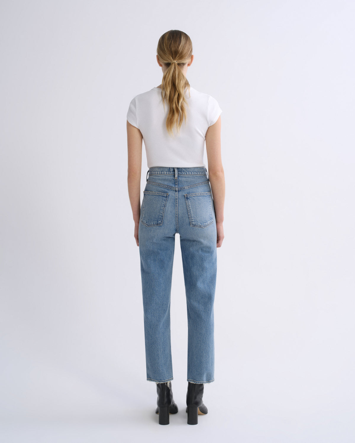 A woman viewed from behind wearing light-wash blue jeans, a white short-sleeve top, and black ankle boots against a plain light gray studio background.