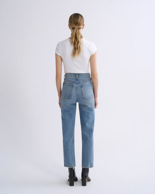 A woman viewed from behind wearing light-wash blue jeans, a white short-sleeve top, and black ankle boots against a plain light gray studio background.