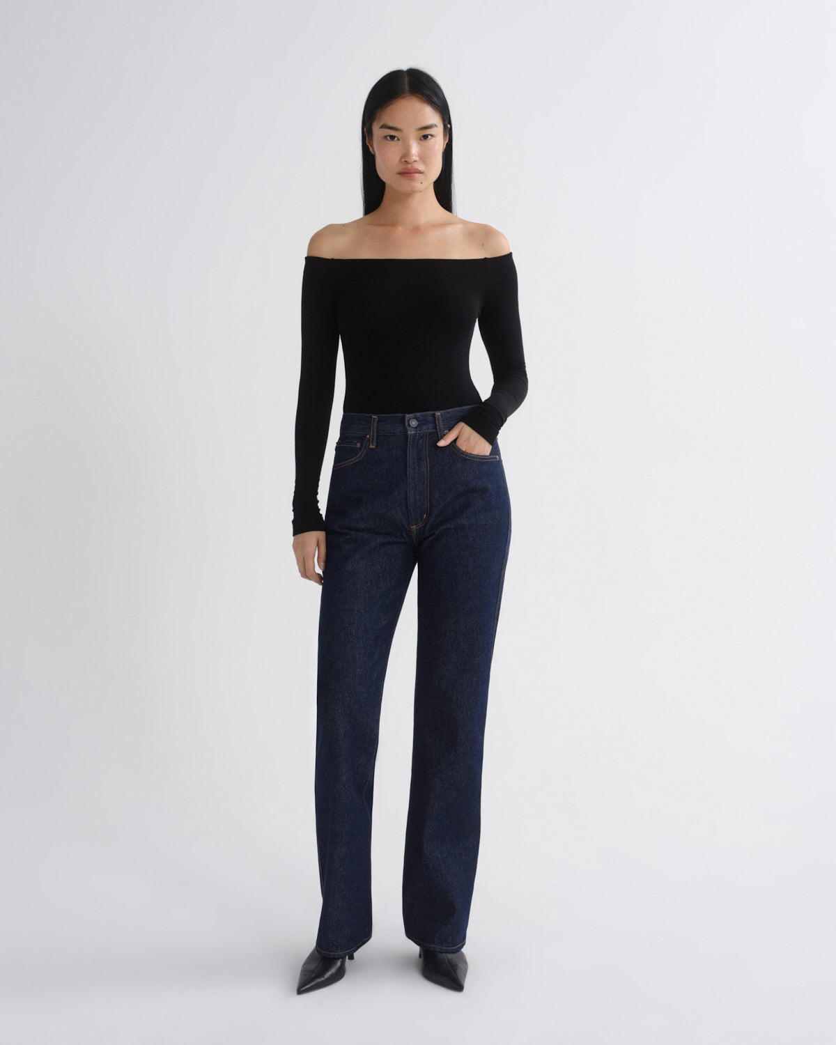 A model stands against a light gray studio background wearing a black off-the-shoulder long-sleeve top and dark blue straight-leg jeans with black pointed shoes.