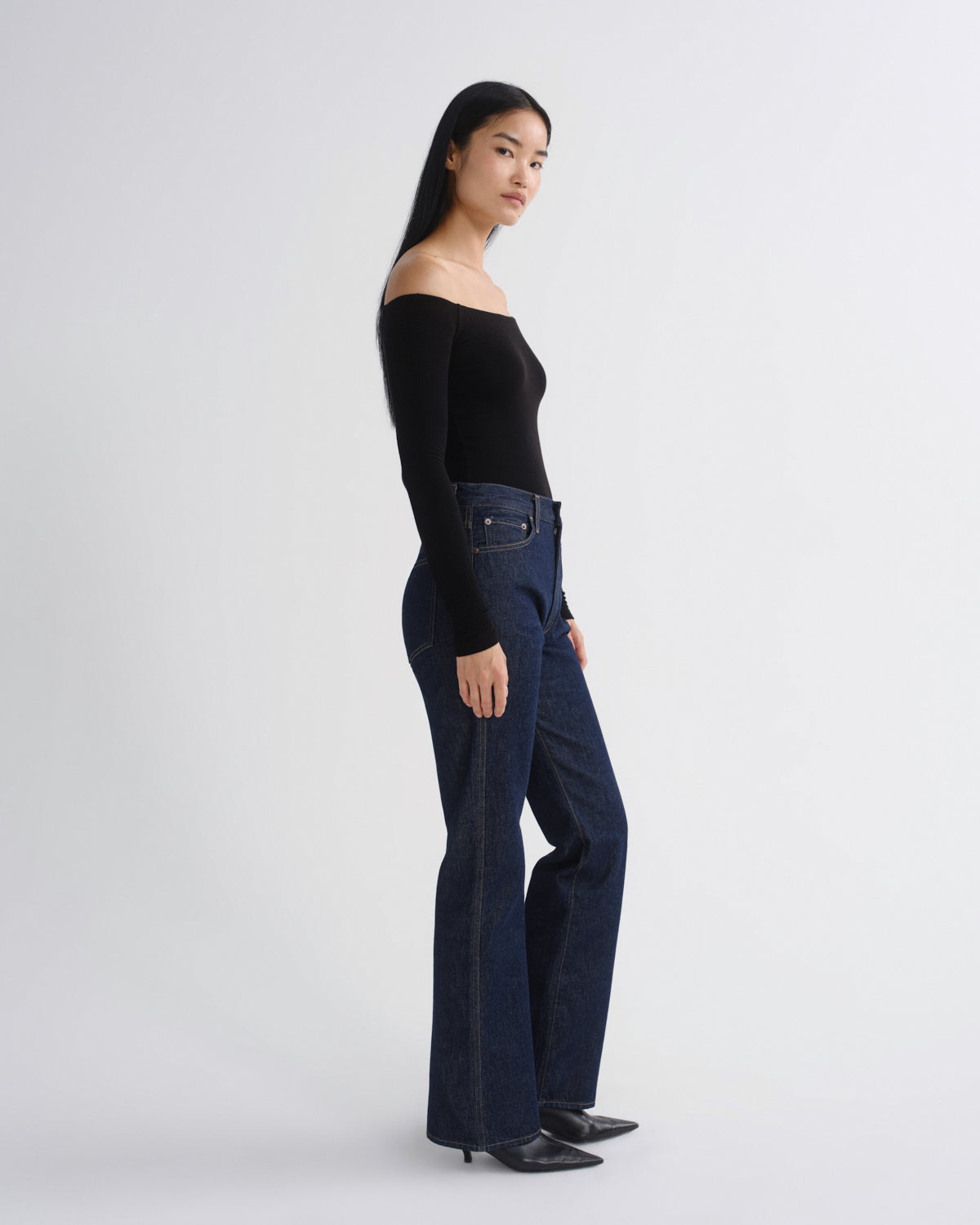 A model is shown in profile wearing a black off-the-shoulder long-sleeve top and dark blue high-waisted flare jeans, standing against a light gray studio background.