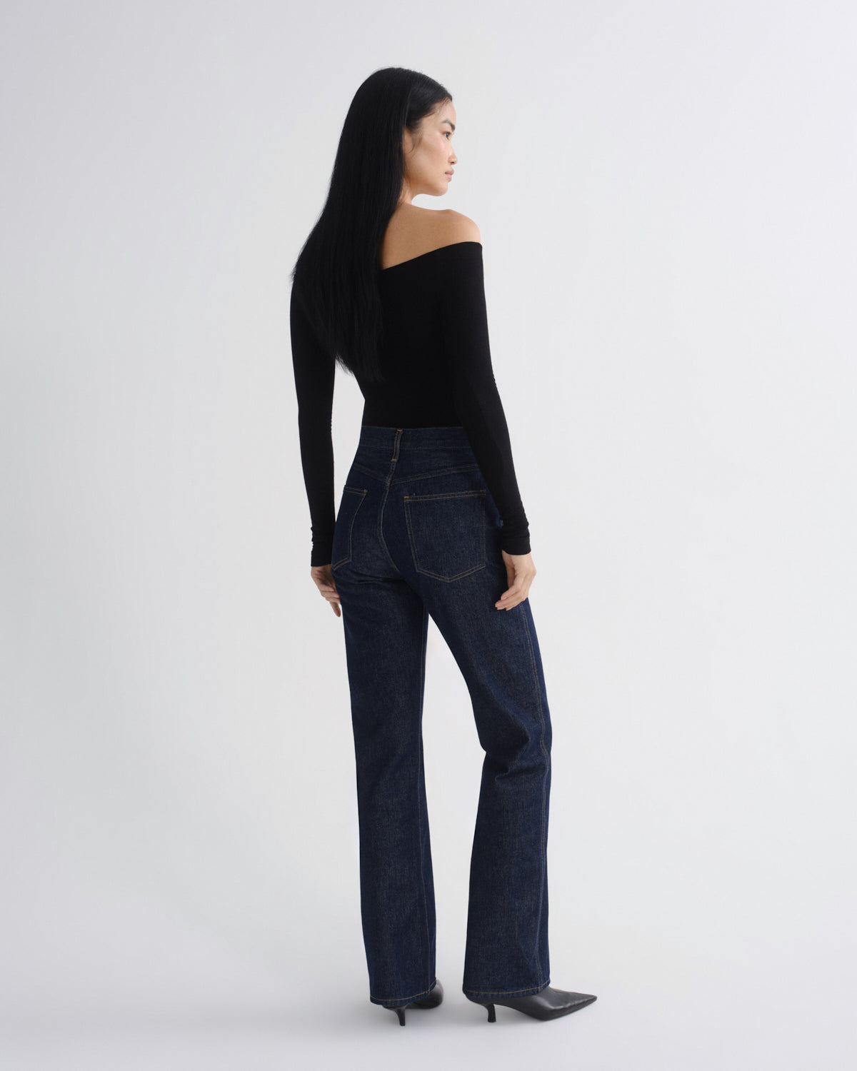 A model shown from the back wearing a black off-the-shoulder long-sleeve top and dark blue high-waisted straight-leg jeans, paired with black pointed-toe heels, against a light gray studio background.