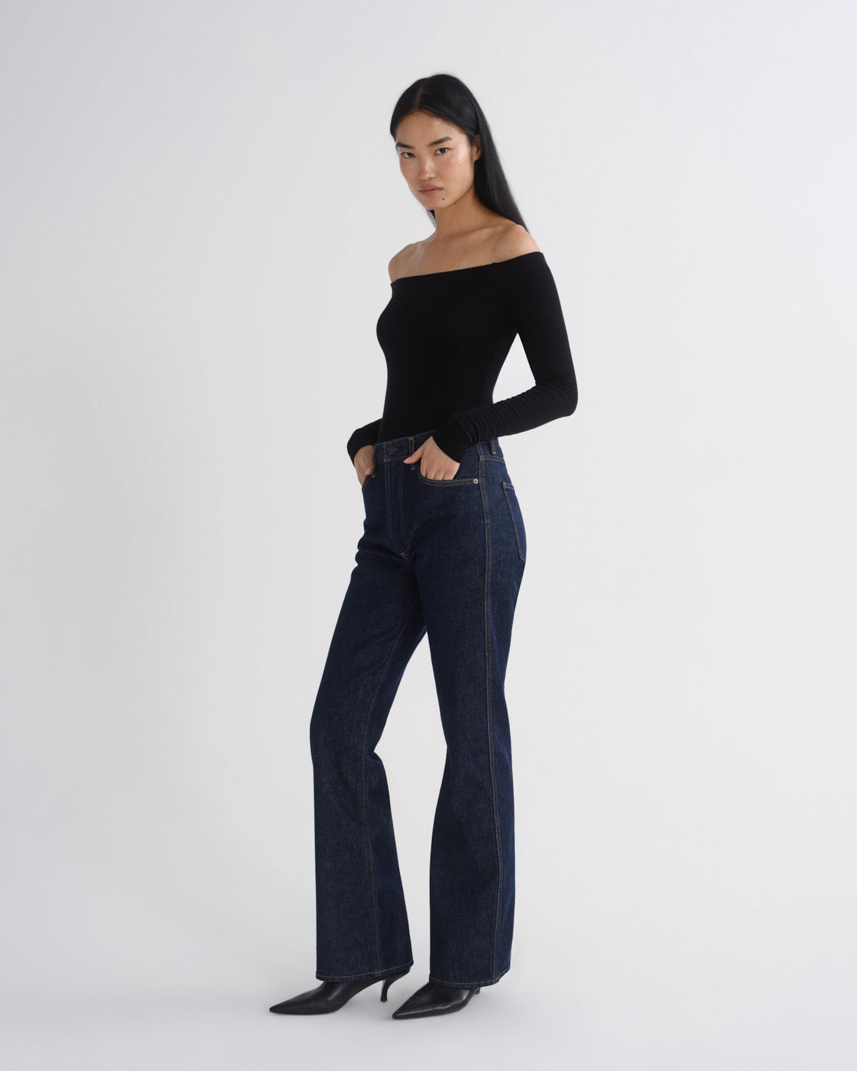 A woman modeling a black off-the-shoulder long-sleeve top and dark blue high-waisted flare jeans with black pointed-toe heels, standing against a light gray studio background.