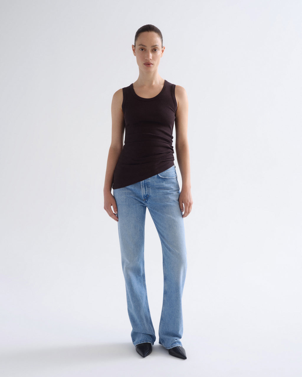 A model stands against a light gray studio background wearing a dark brown sleeveless top, light blue jeans, and black pointed shoes.