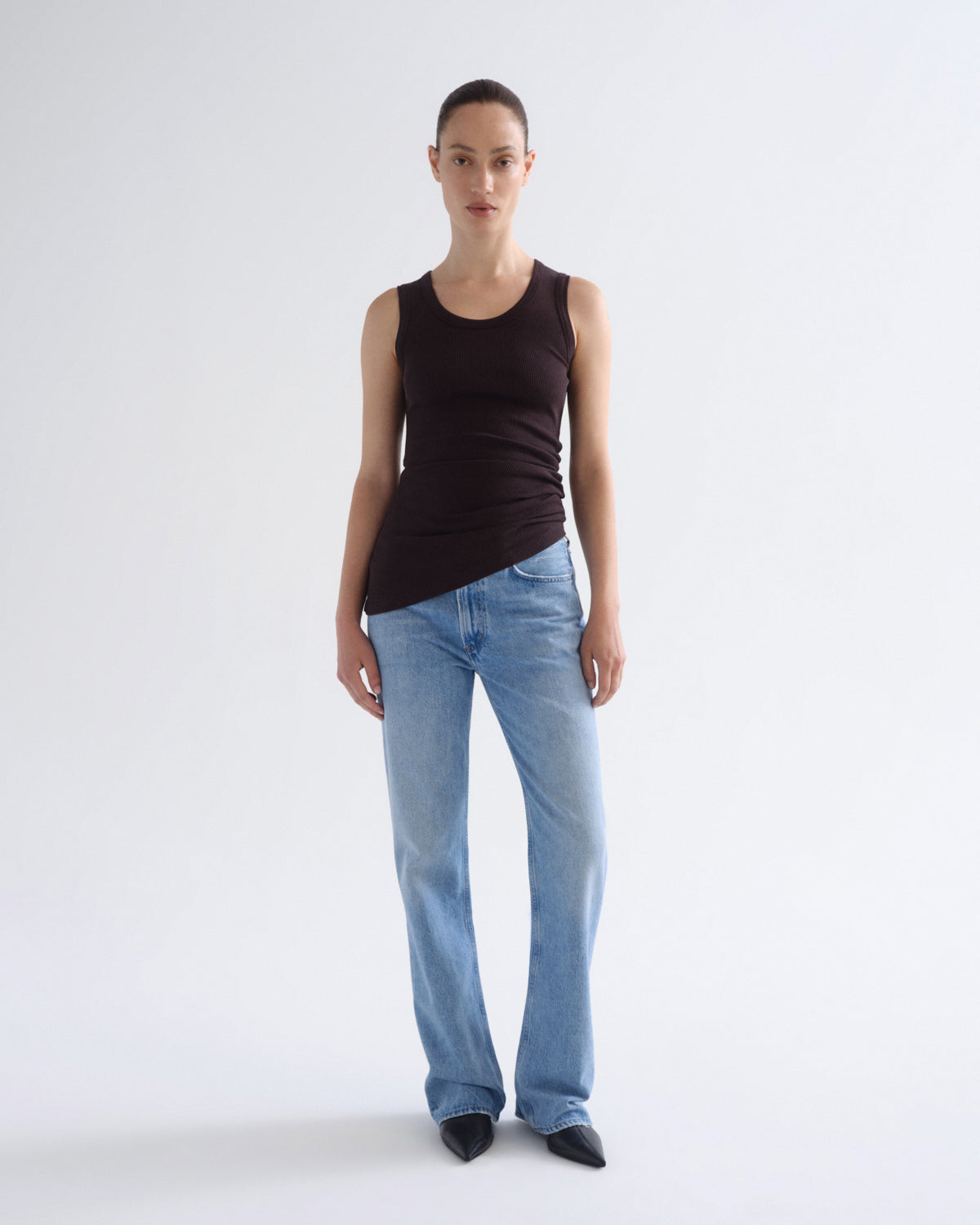 A model stands against a light gray studio background wearing a dark brown sleeveless top, light blue jeans, and black pointed shoes.