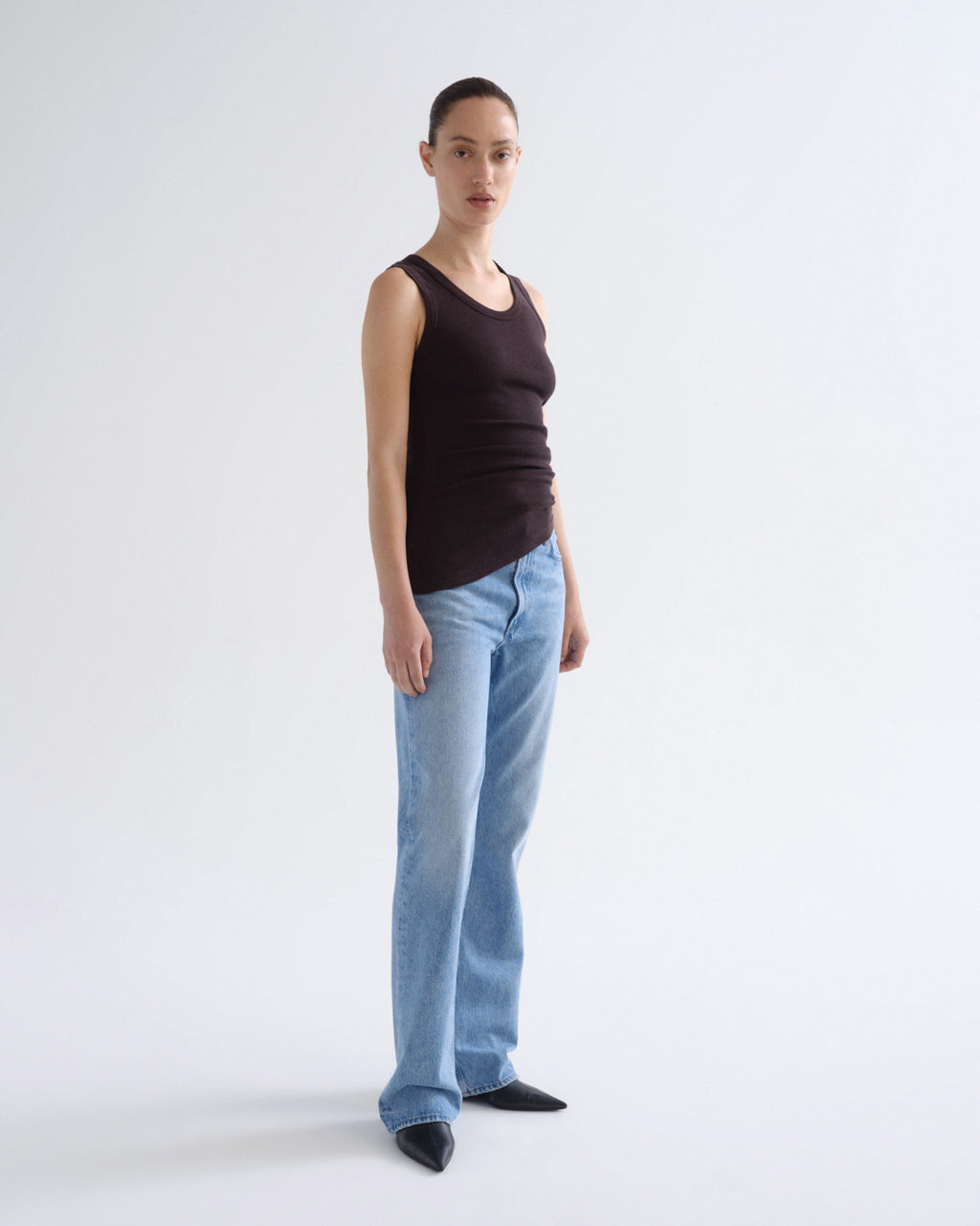 A model stands in profile wearing a dark brown ribbed tank top, light blue straight-leg jeans, and black pointed shoes against a plain light gray studio background.