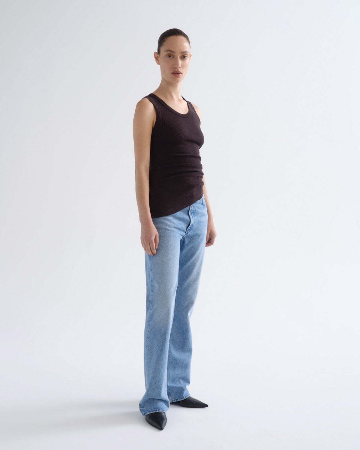 A model stands in profile wearing a dark brown ribbed tank top, light blue straight-leg jeans, and black pointed shoes against a plain light gray studio background.
