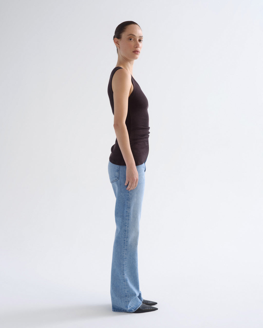 A woman stands in profile wearing a dark brown sleeveless top and light blue flared jeans against a plain white studio background.