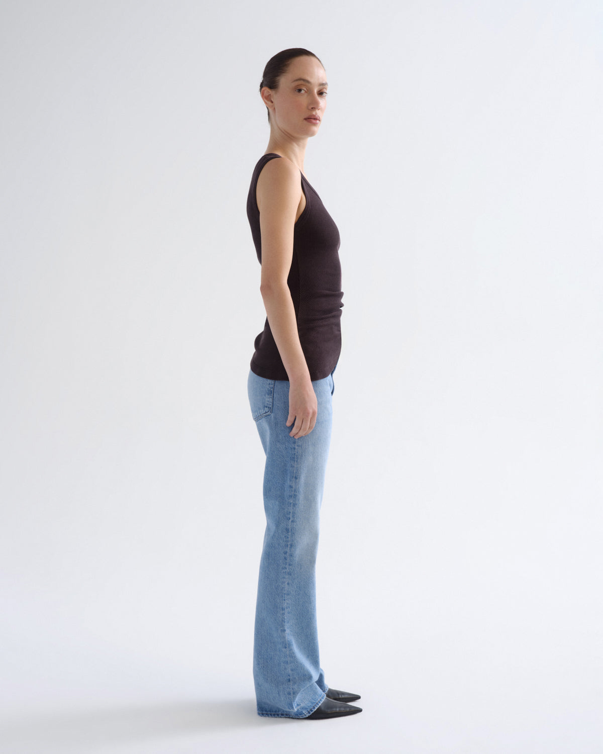 A woman stands in profile wearing a dark brown sleeveless top and light blue flared jeans against a plain white studio background.