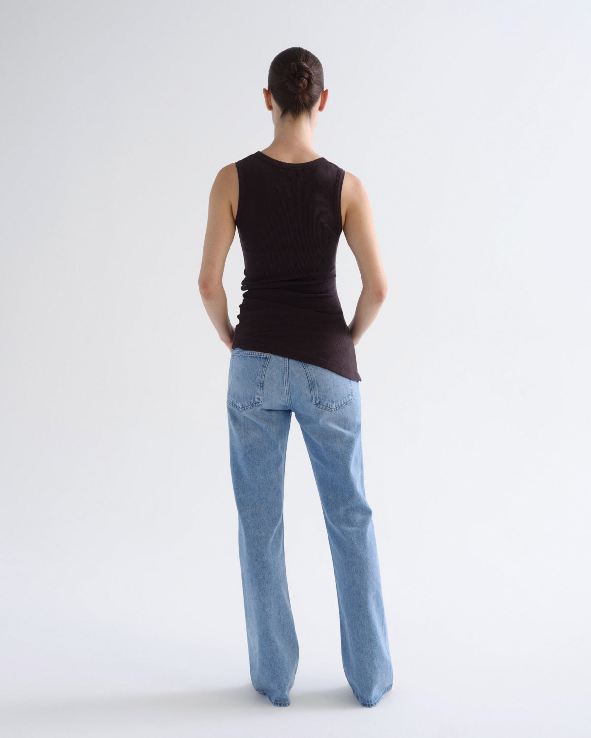 A model viewed from behind wearing a dark brown sleeveless top and light blue jeans against a plain white studio background.