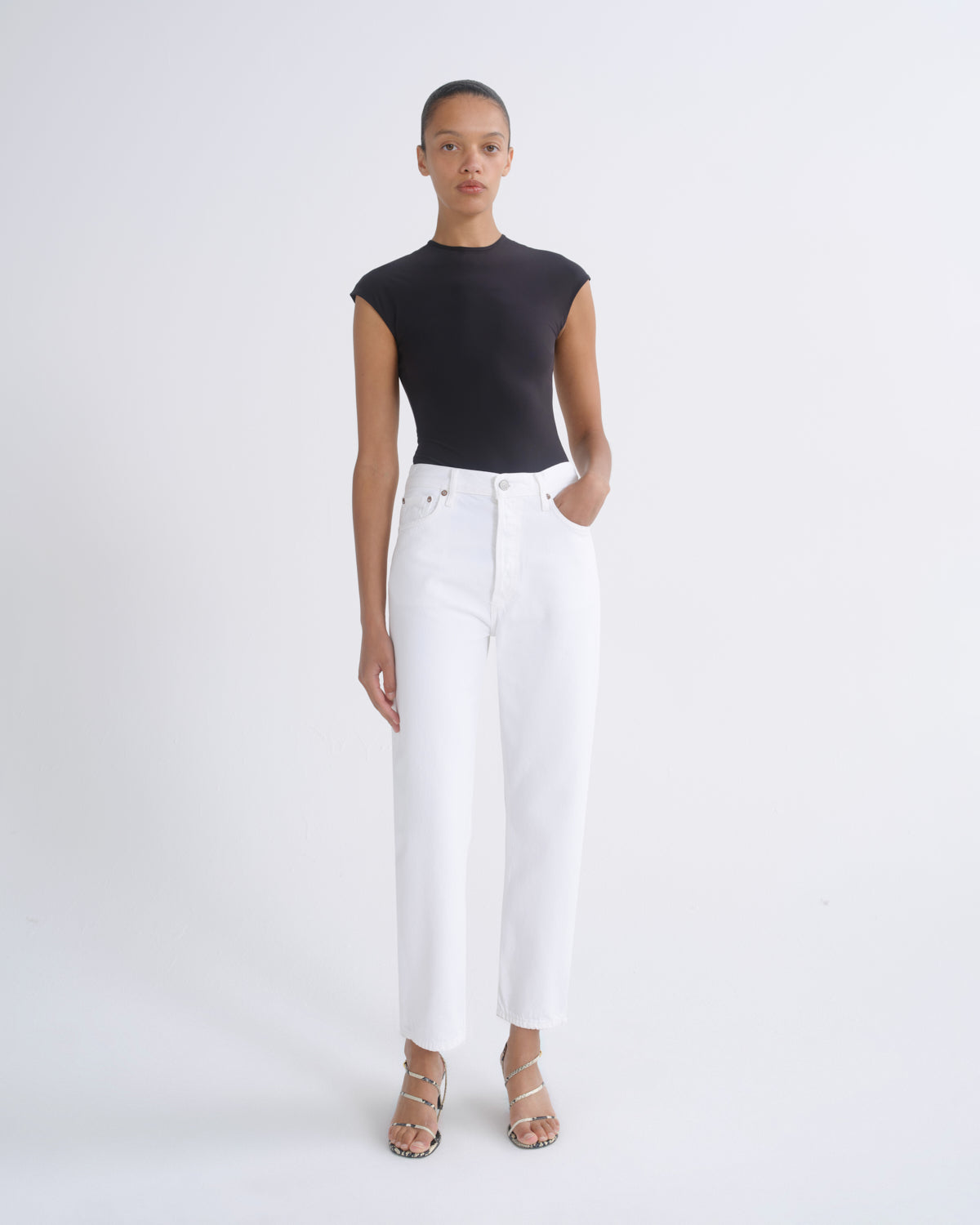 A model stands against a plain white studio background wearing a black sleeveless mock-neck top, white wide-leg cropped jeans, and beige strappy sandals.
