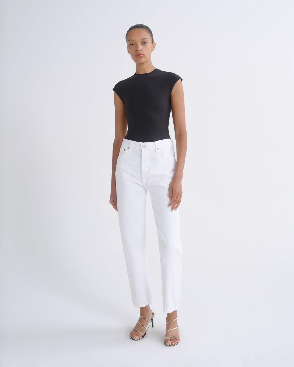 A model stands against a light gray studio background wearing a black sleeveless top and white high-waisted cropped jeans with metallic sandals.