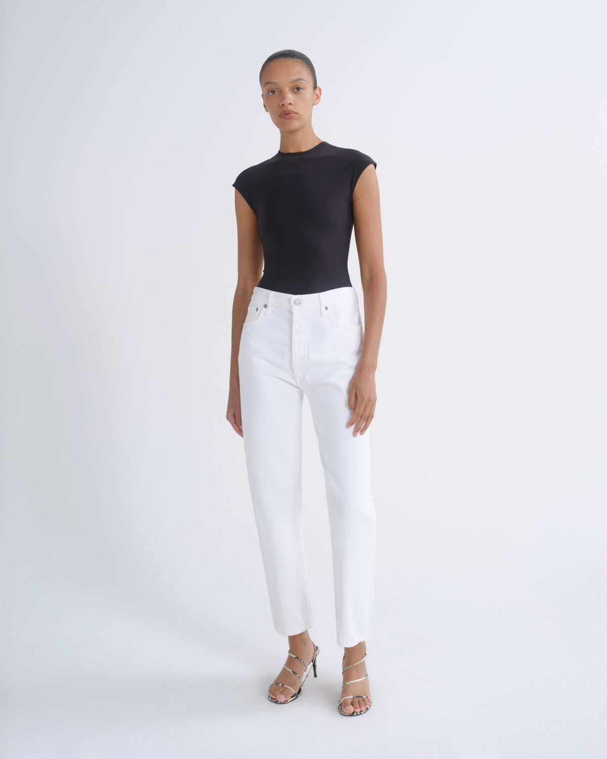 A model stands against a light gray studio background wearing a black sleeveless top and white high-waisted cropped jeans with metallic sandals.