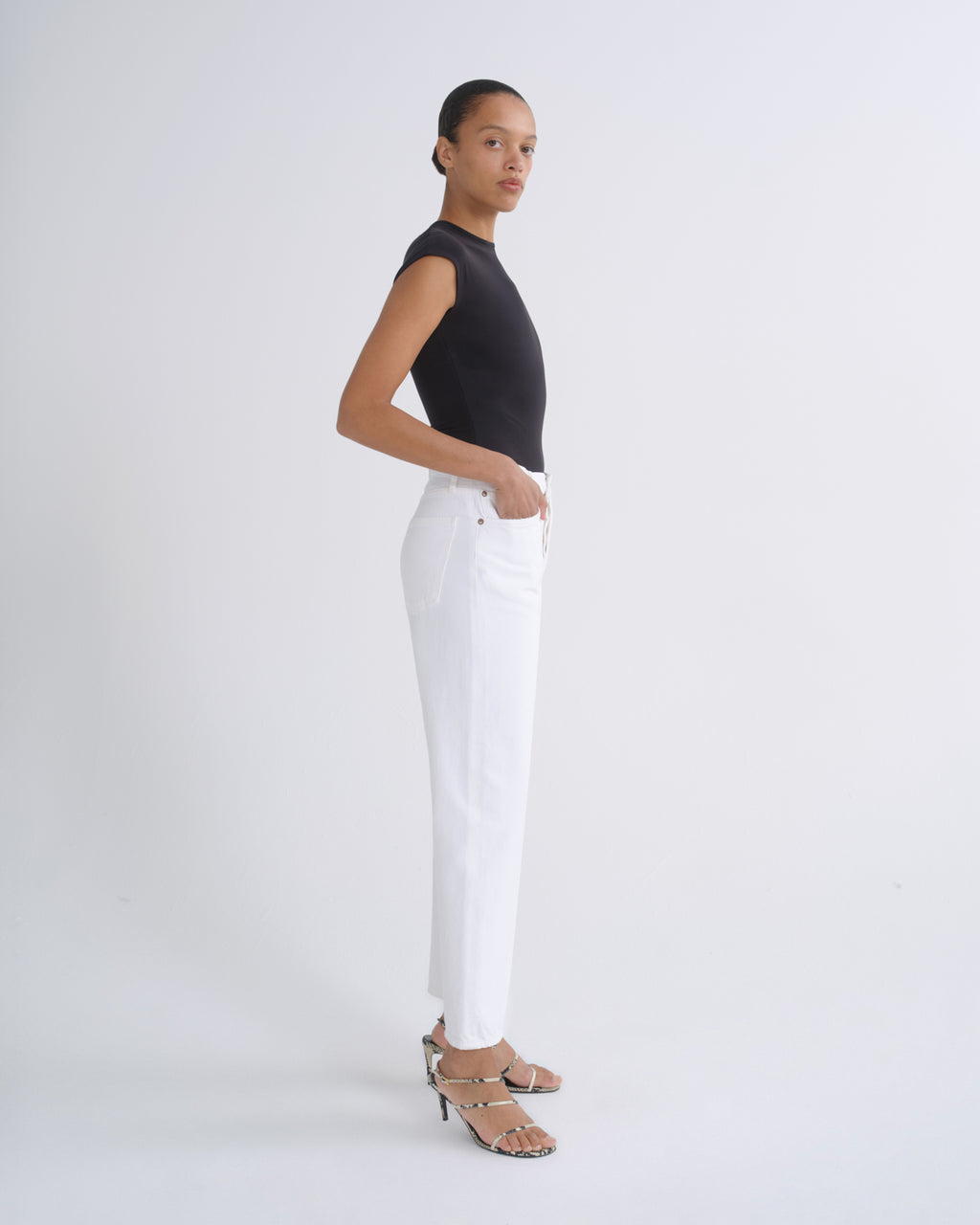 A model stands in profile wearing a black sleeveless top, white straight-leg pants, and strappy high-heeled sandals against a clean white studio background.