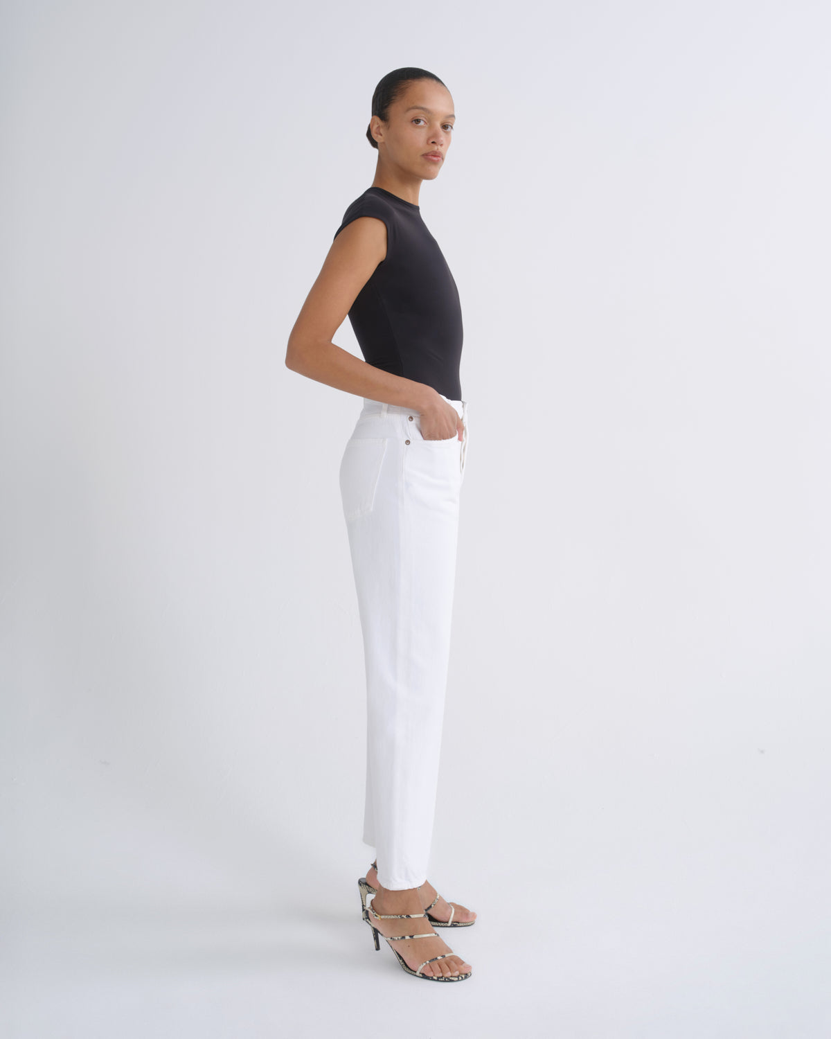 A model stands in profile wearing a black sleeveless top, white straight-leg pants, and strappy high-heeled sandals against a clean white studio background.