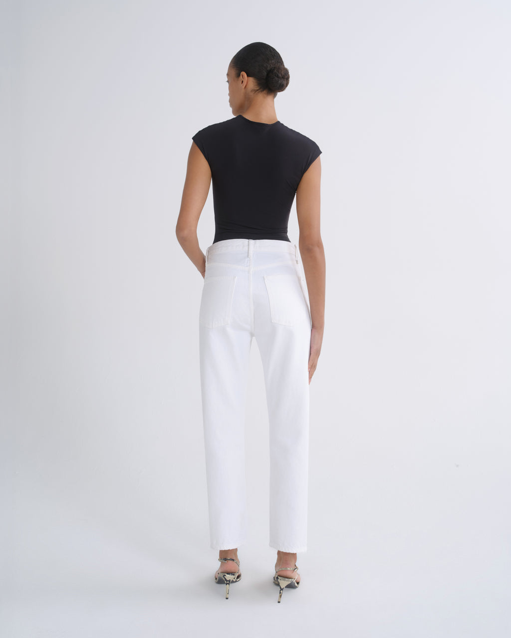 A woman is shown from the back wearing a black sleeveless top and white straight-leg trousers, paired with metallic high-heeled sandals, against a plain light gray studio background.