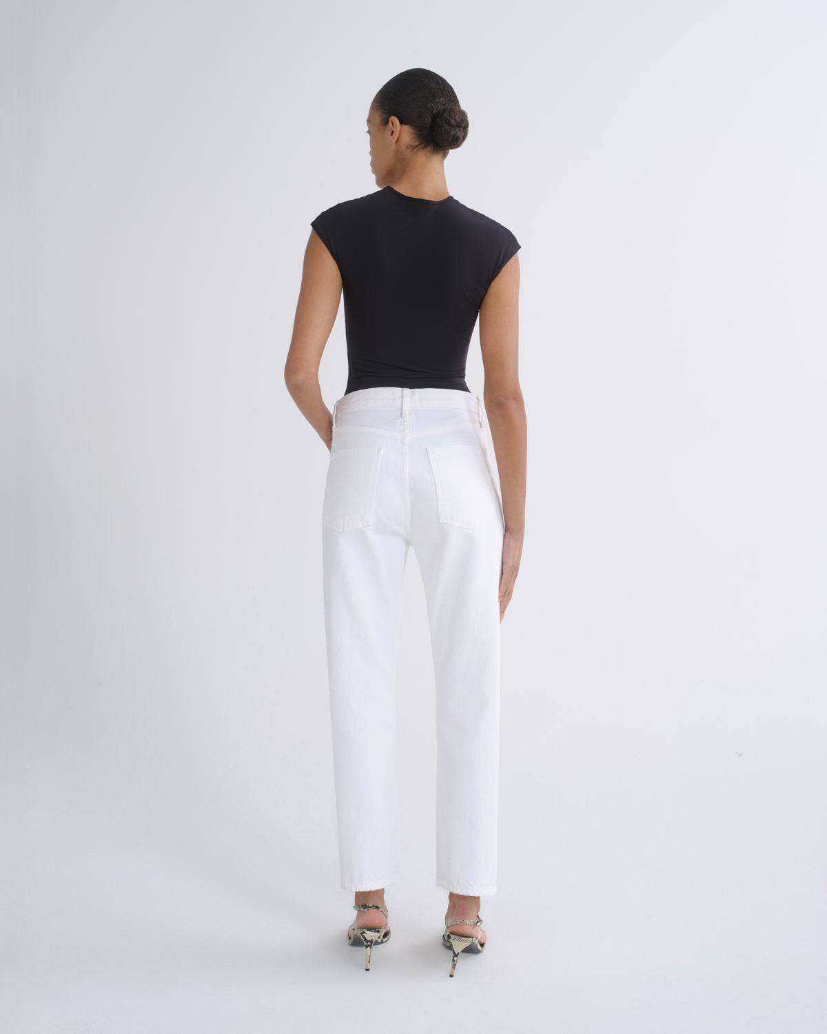 A woman is shown from the back wearing a black sleeveless top and white straight-leg trousers, paired with metallic high-heeled sandals, against a plain light gray studio background.