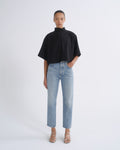 A model stands against a light gray studio background wearing a black short-sleeve mock-neck top, light-wash blue straight-leg jeans, and beige strappy high-heeled sandals.