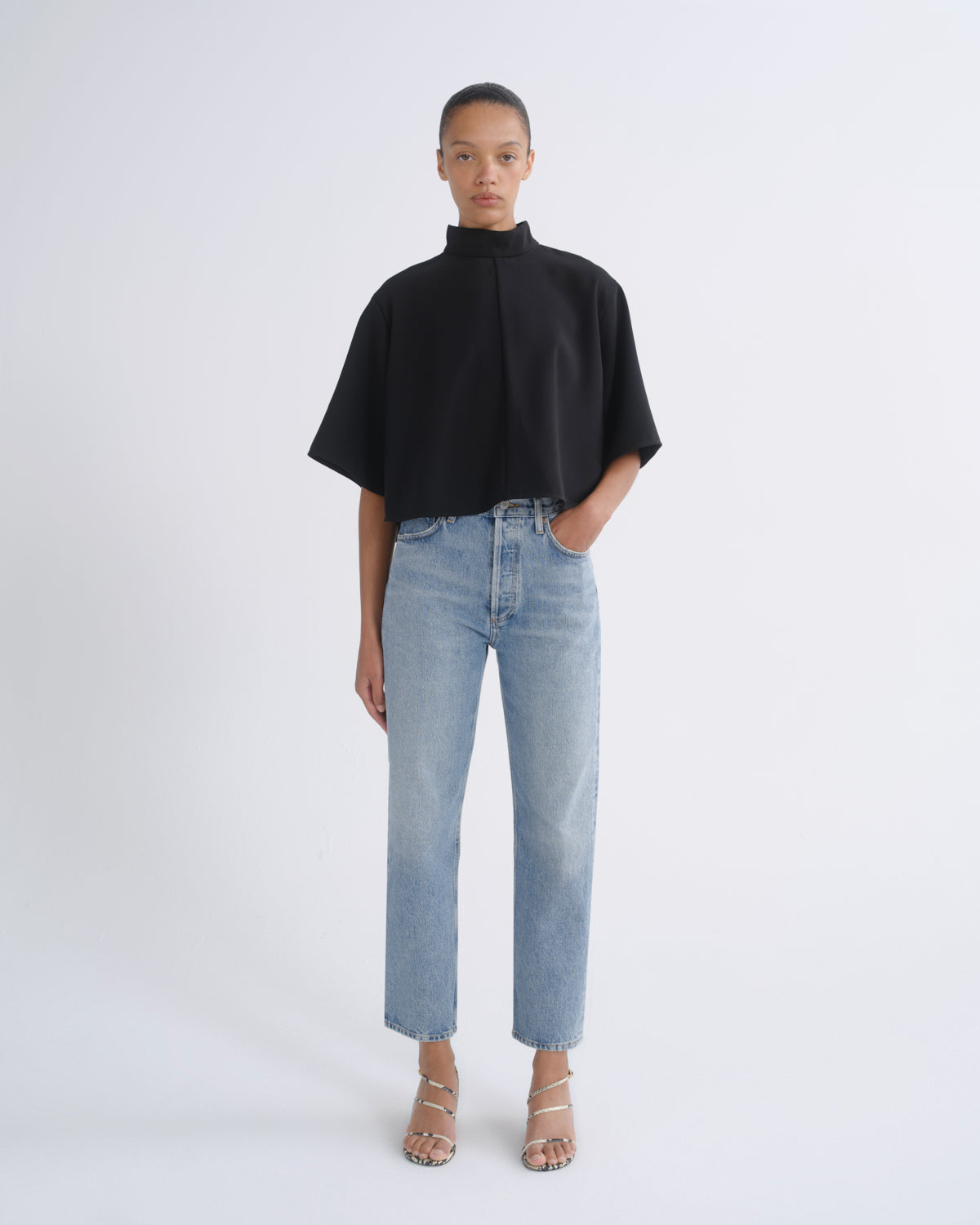 A model stands against a light gray studio background wearing a black short-sleeve mock-neck top, light-wash blue straight-leg jeans, and beige strappy high-heeled sandals.