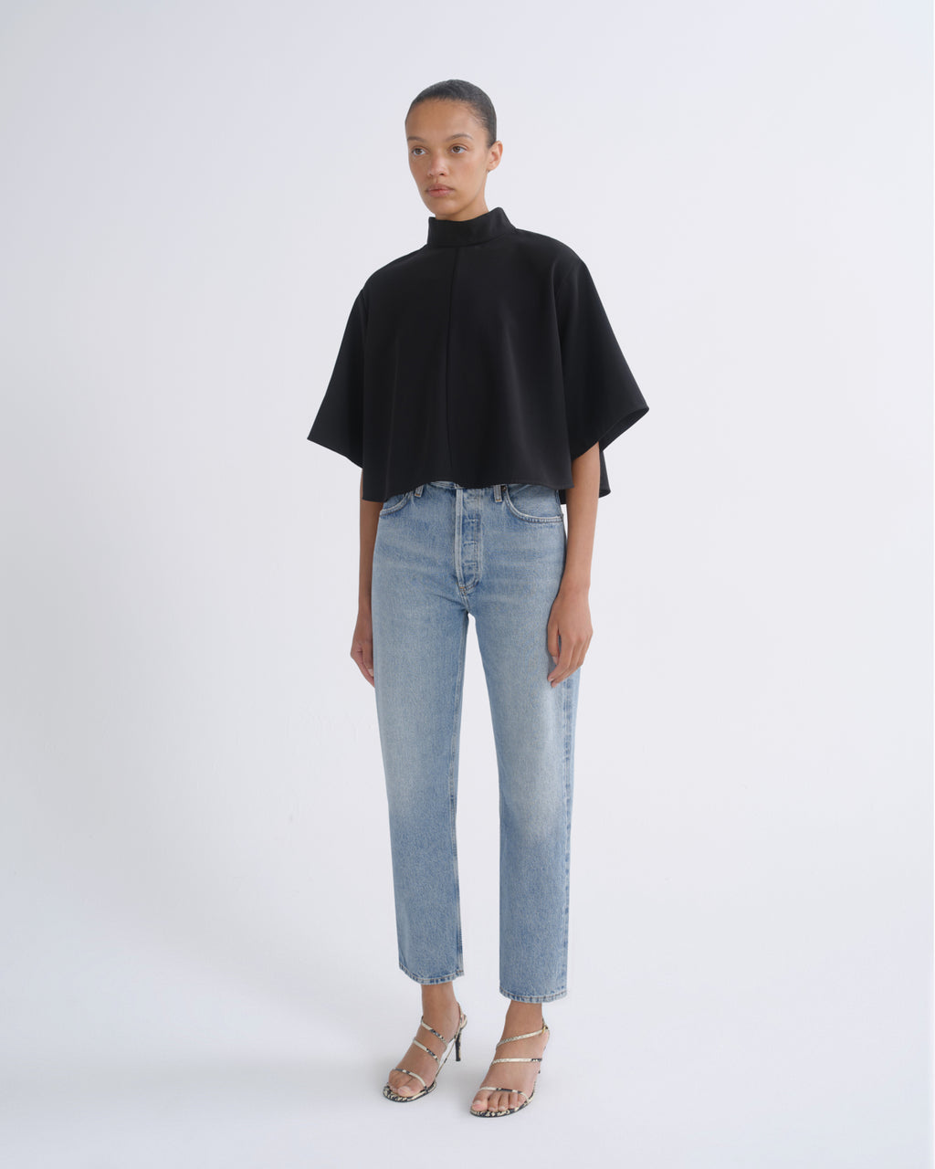 A model stands against a light gray studio background wearing a black high-neck cape-sleeve top, light-wash blue jeans, and metallic heeled sandals.