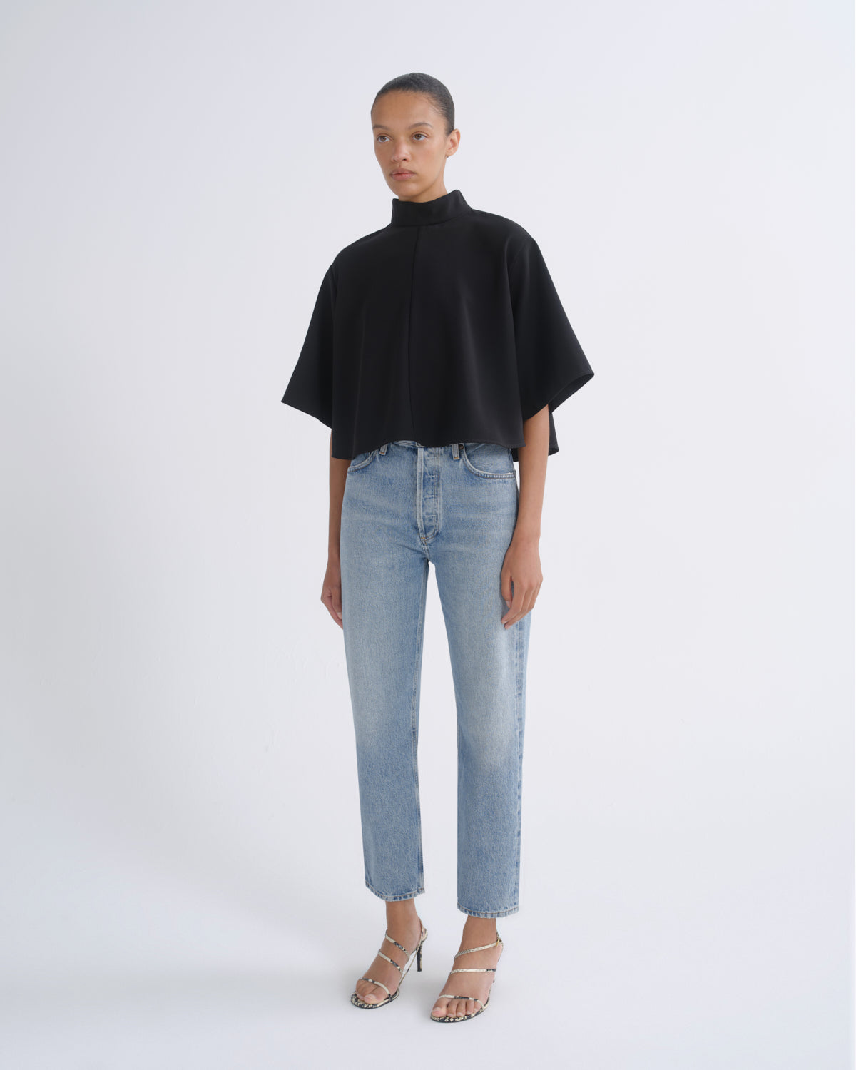 A model stands against a light gray studio background wearing a black high-neck cape-sleeve top, light-wash blue jeans, and metallic heeled sandals.