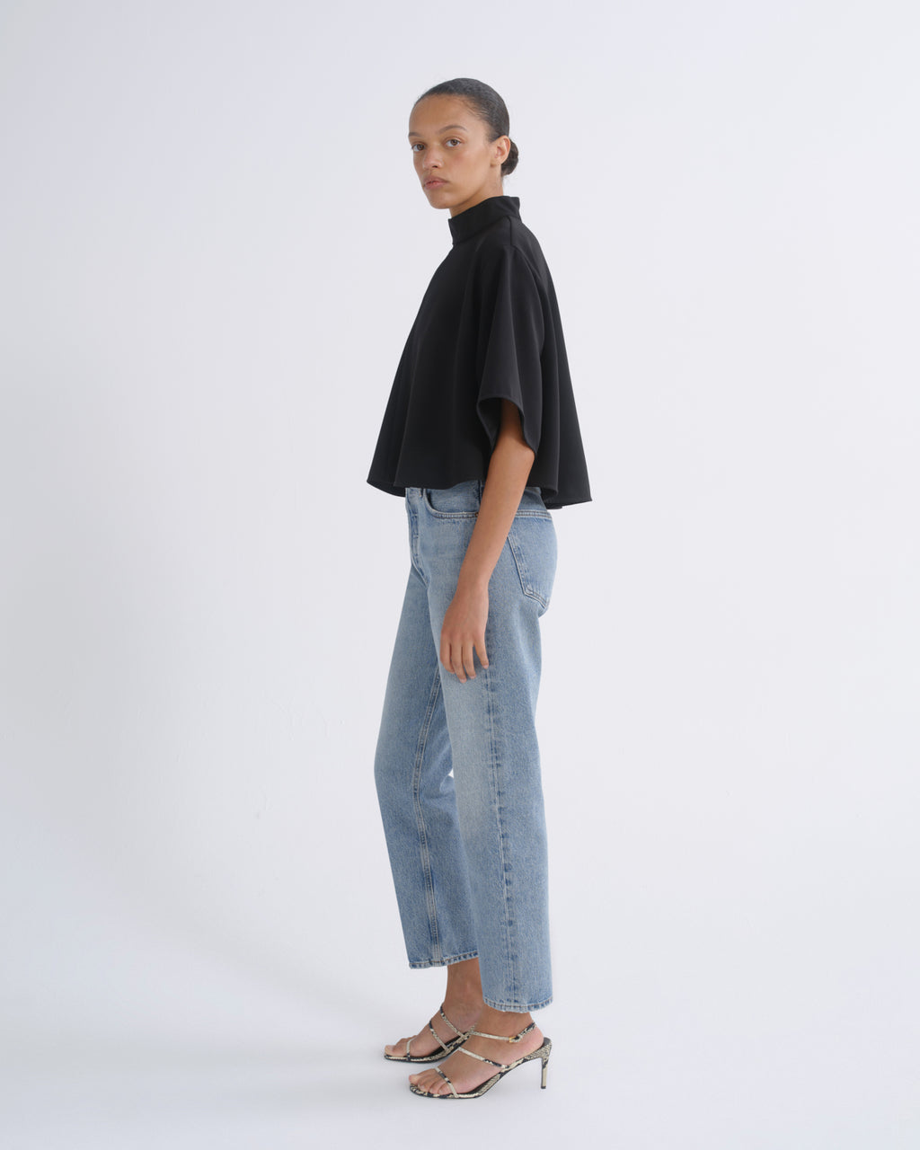 A model is shown in profile wearing a black short-sleeved mock-neck top, light-wash blue cropped jeans, and metallic silver heeled sandals against a plain white studio background.