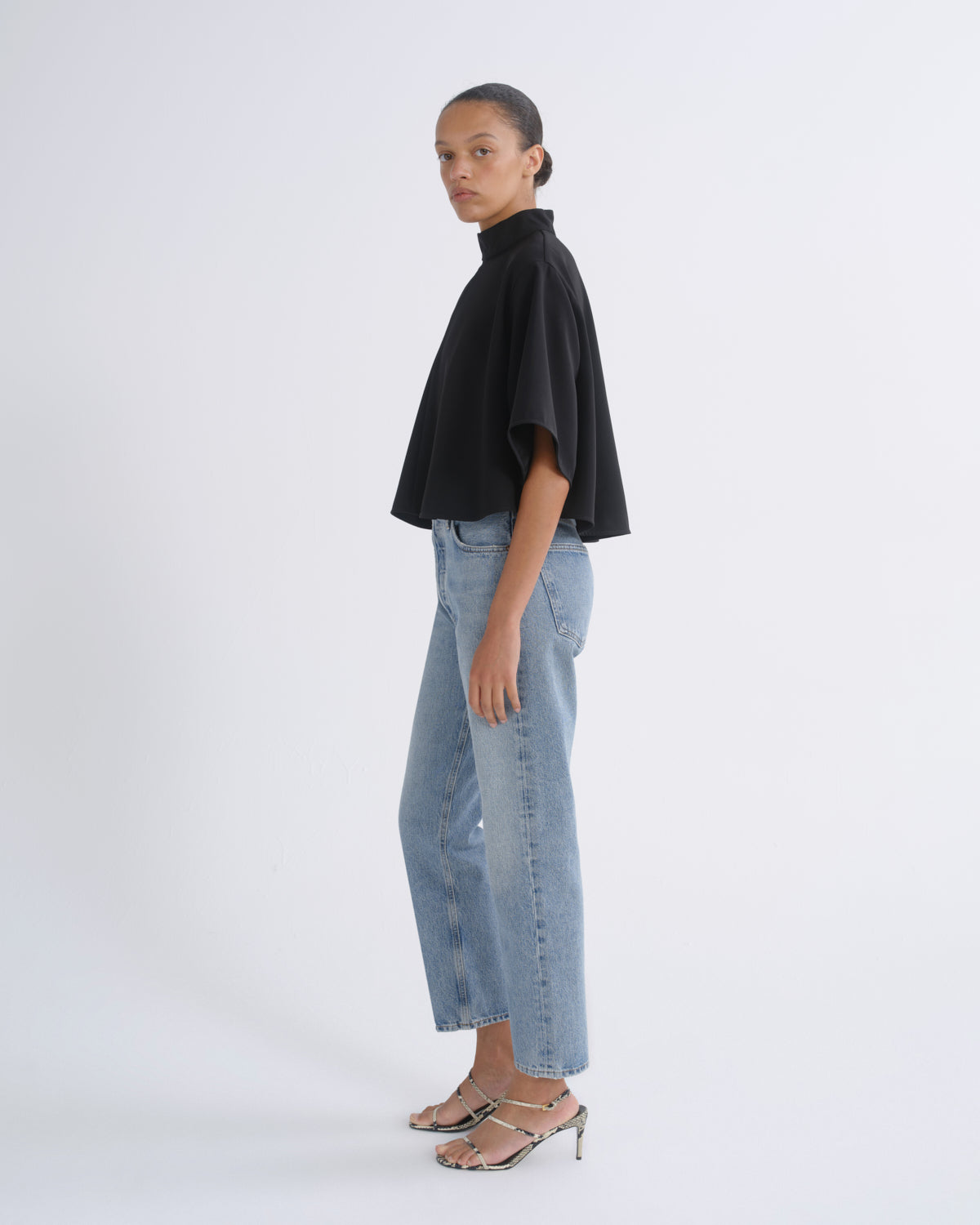 A model is shown in profile wearing a black short-sleeved mock-neck top, light-wash blue cropped jeans, and metallic silver heeled sandals against a plain white studio background.