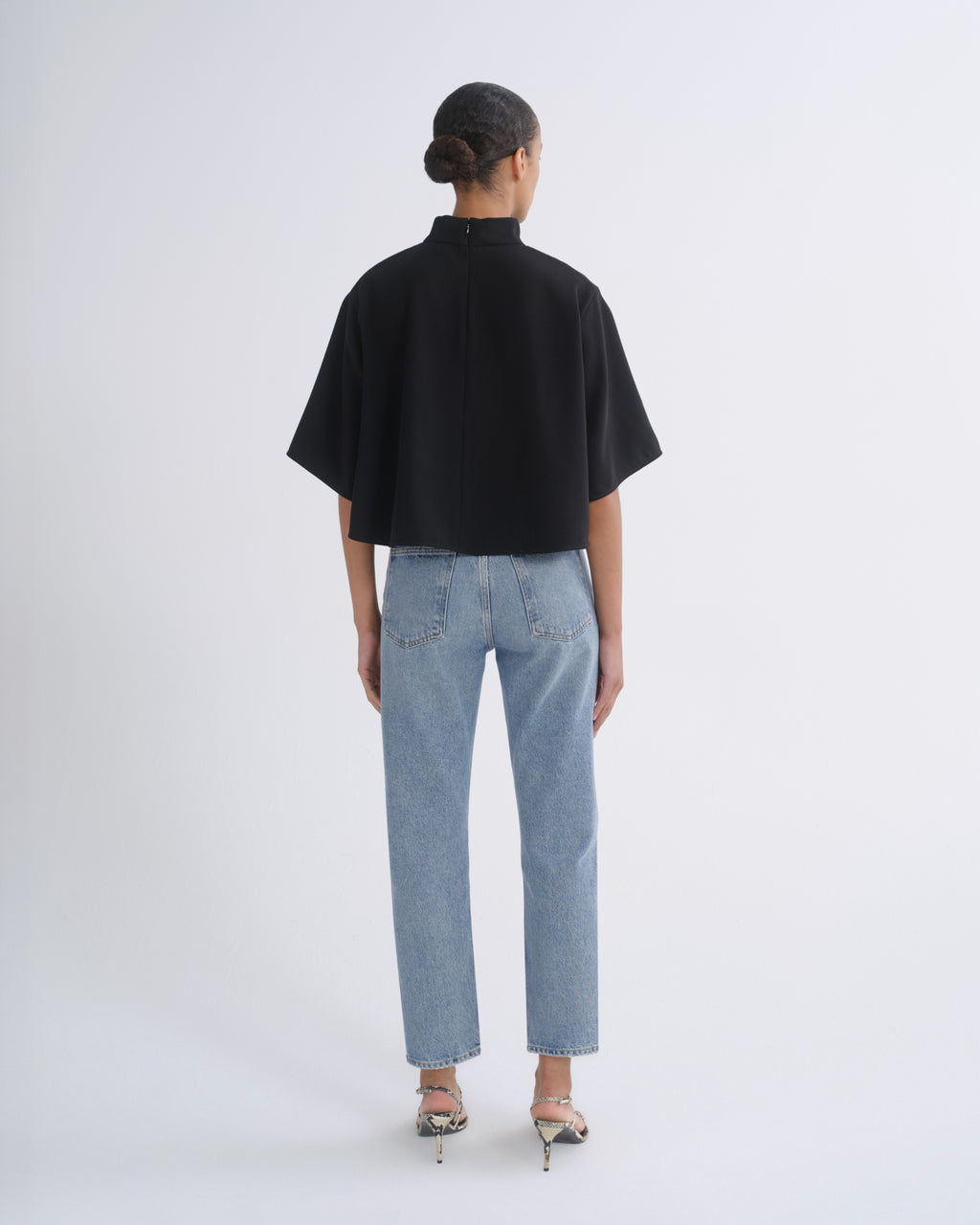 A model viewed from the back wearing a black short-sleeved mock-neck top and light-wash blue jeans, standing against a plain light gray studio background.