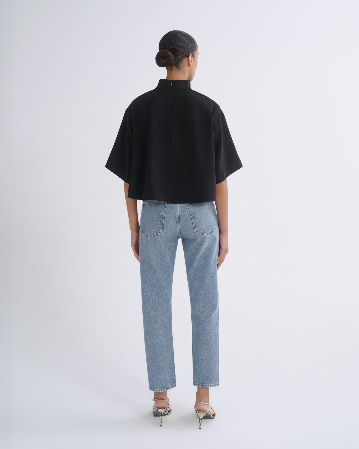 A model viewed from the back wearing a black short-sleeved mock-neck top and light-wash blue jeans, standing against a plain light gray studio background.