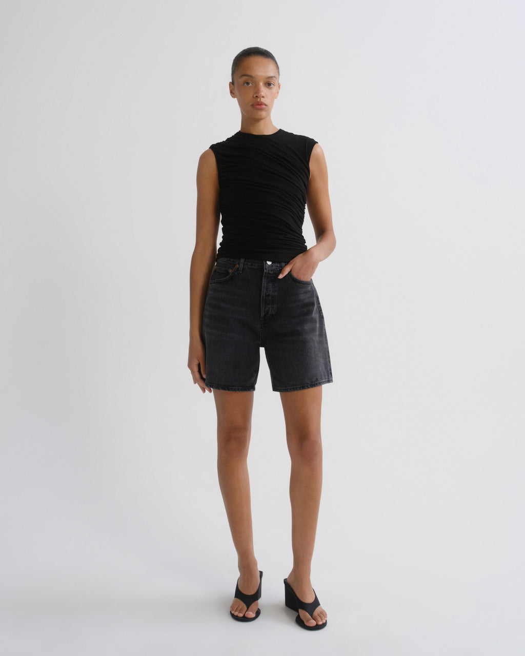 A model stands against a light gray studio background wearing a sleeveless black ruched top, dark denim shorts, and black sandals.