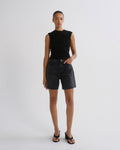 A model stands against a light gray studio background wearing a sleeveless black ruched top, dark denim shorts, and black sandals.