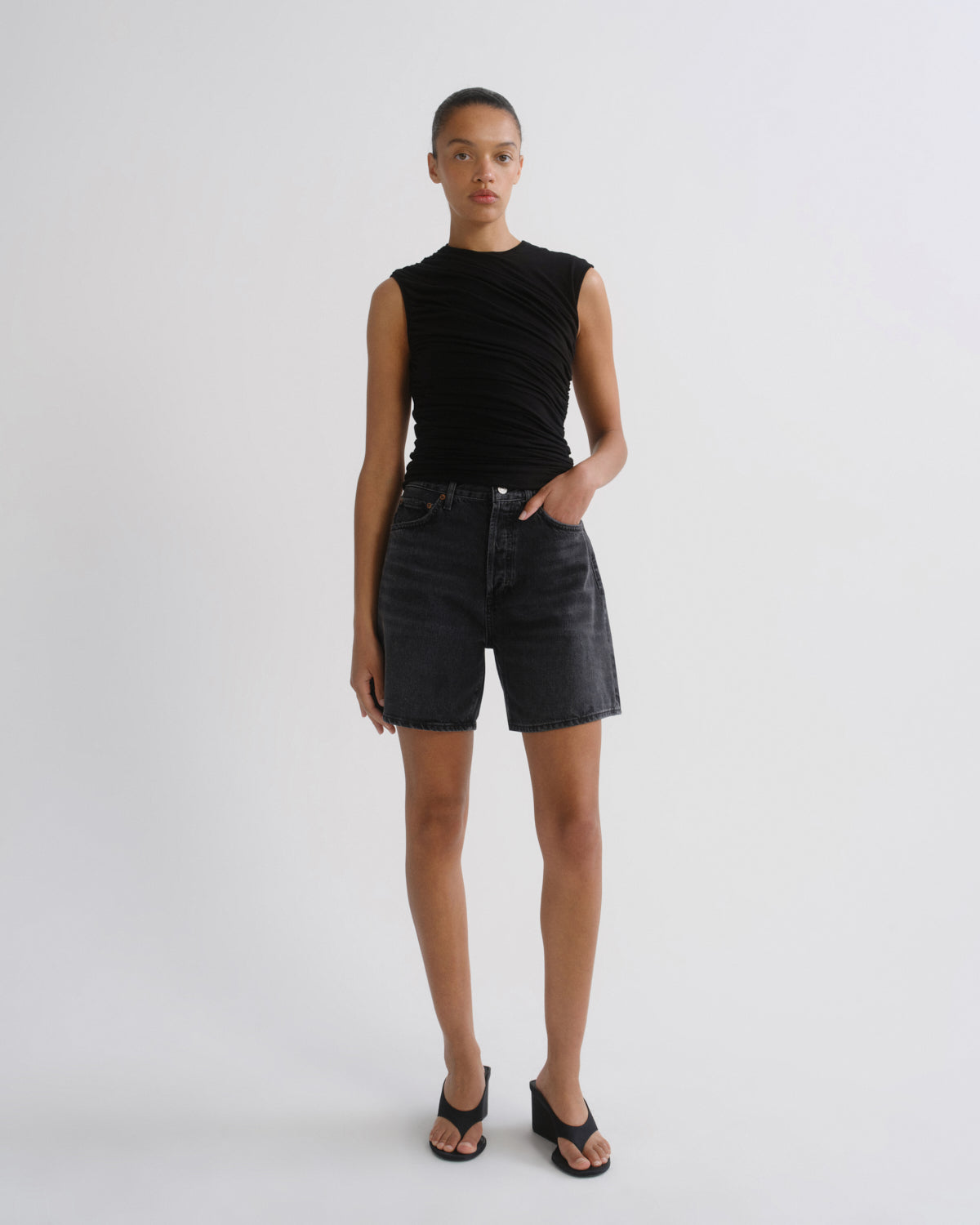 A model stands against a light gray studio background wearing a sleeveless black ruched top, dark denim shorts, and black sandals.